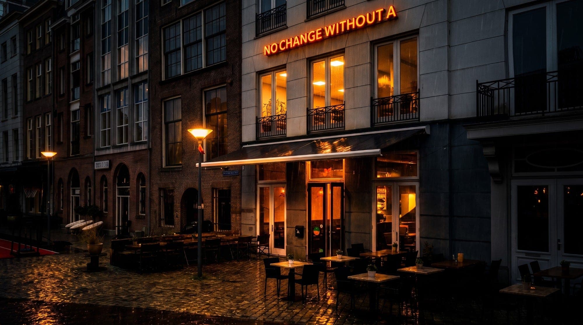 Het pand aan de Oost-Wijnstraat op een regenachtige avond, met warm licht achter de ramen en een nat terras
